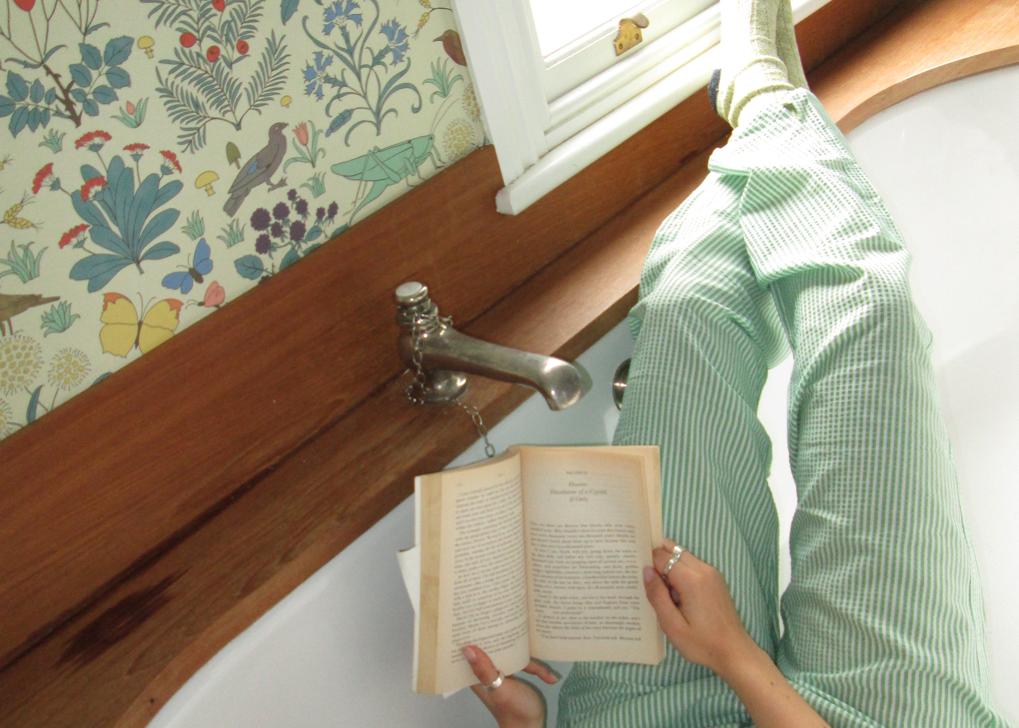 Relaxing in green striped lounge pants, reading a book by the window with floral wallpaper.
