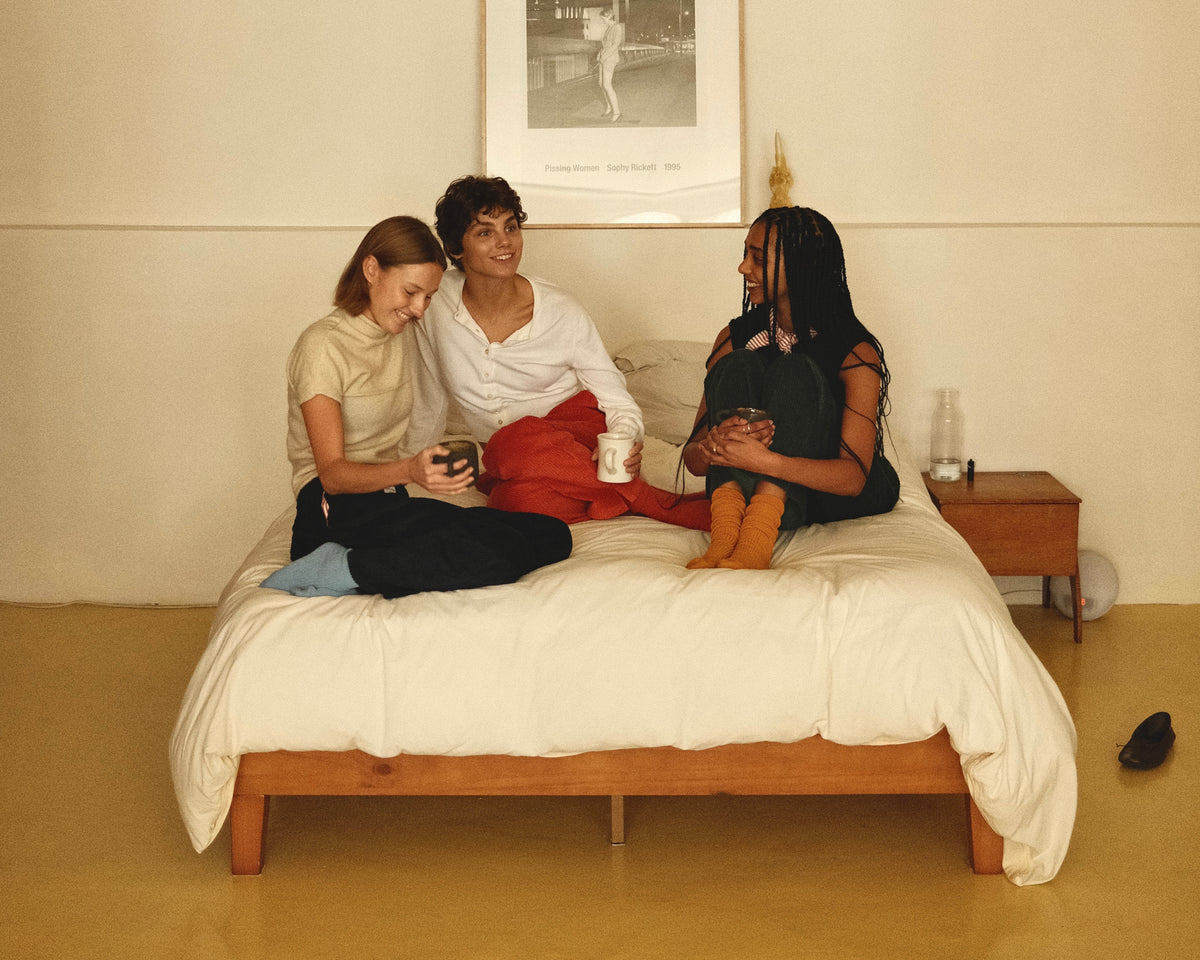 Three people sitting together on a bed wearing Speltham corduroy lounge trousers, with a wooden bed frame, white bedding, a bedside table, and a framed print on the wall.