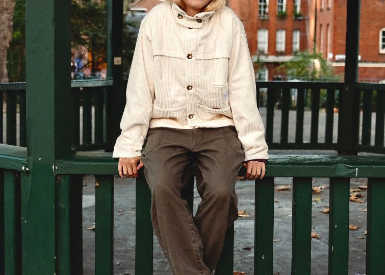 Person in the cream corduroy jacket and a cap sitting on a green railing in a London square