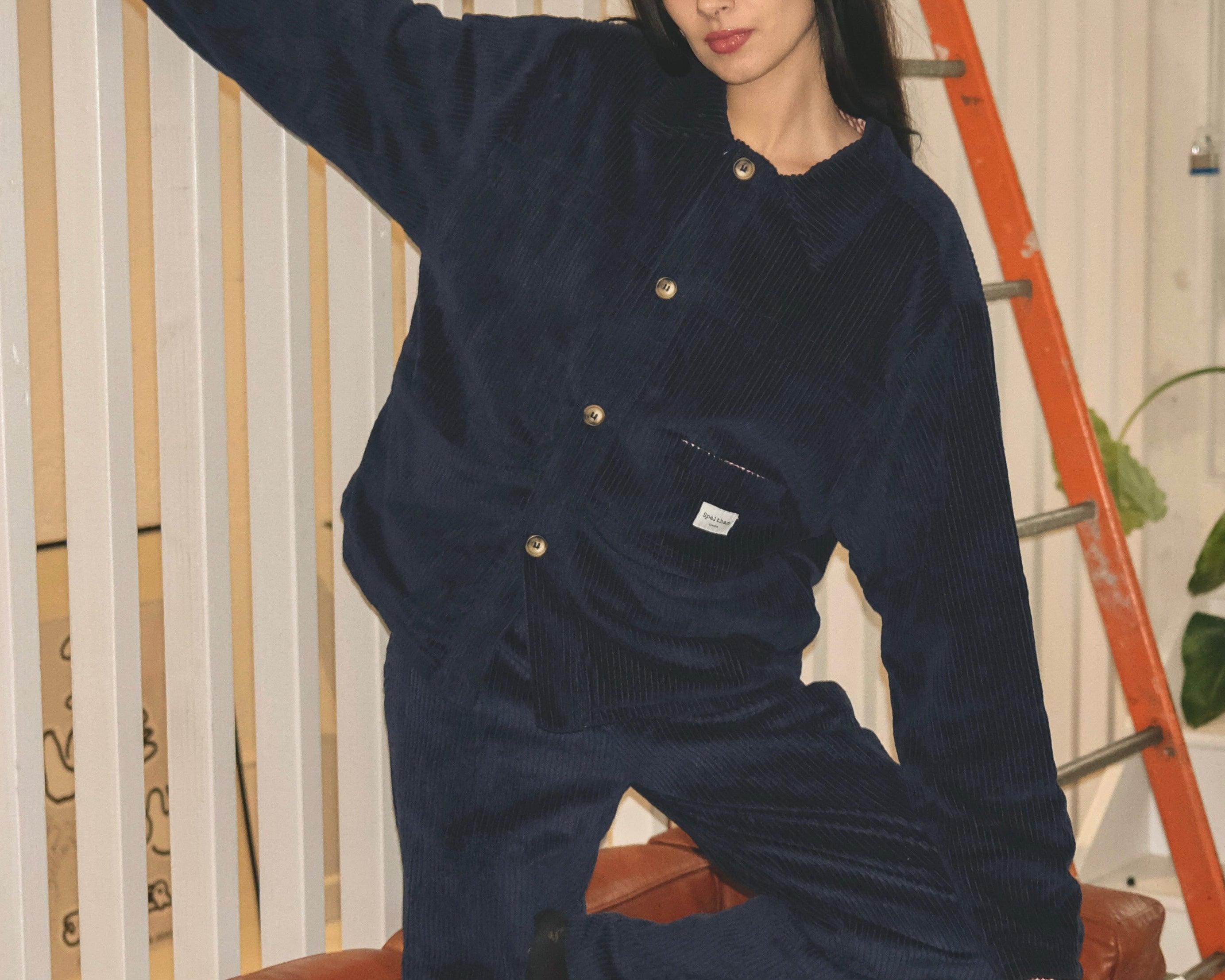 Woman in the navy corduroy jacket and matching trousers, one arm raised, sitting on an orange chair