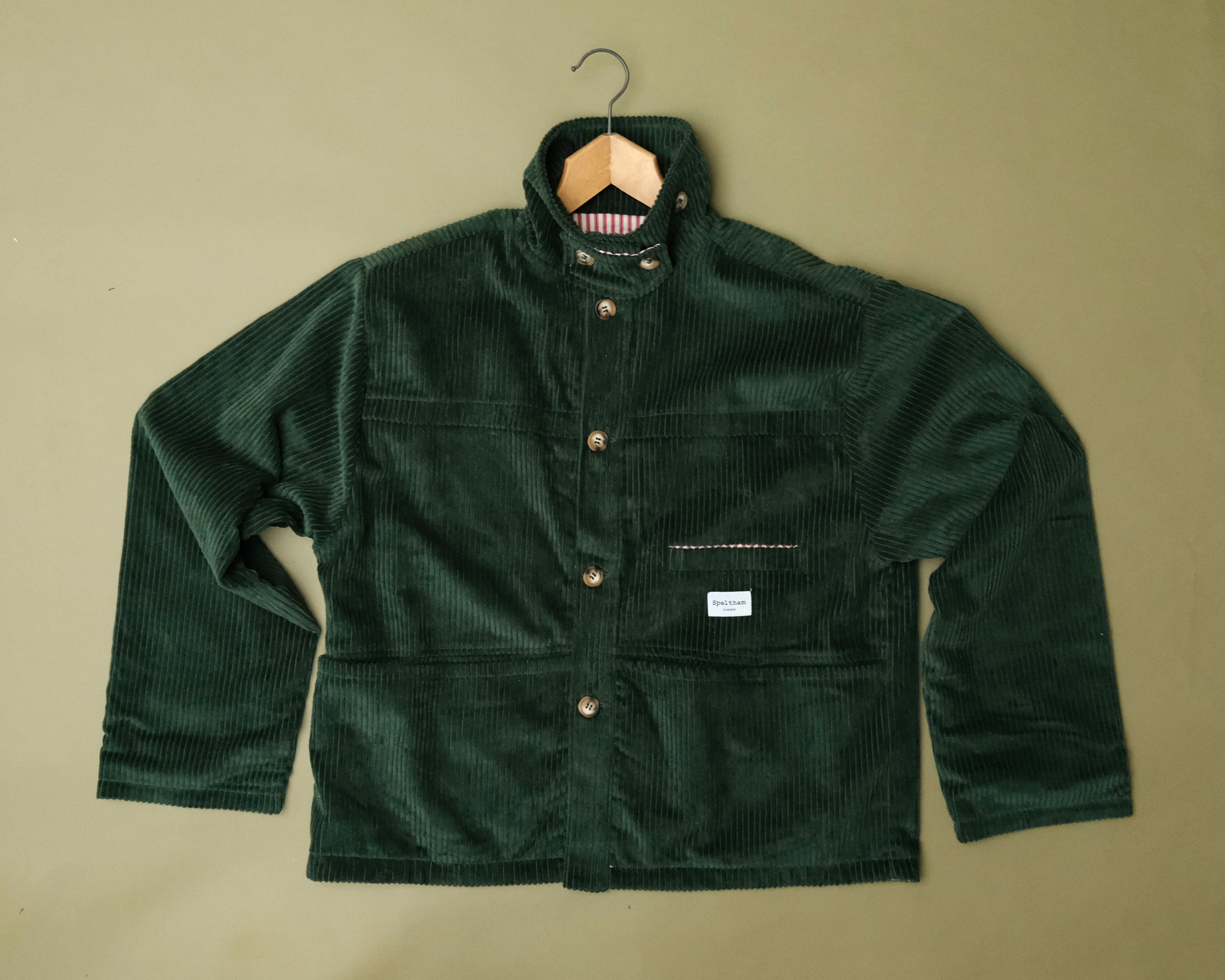 Speltham green corduroy jacket on a hanger against a sage green wall, showing the button front and patch pockets