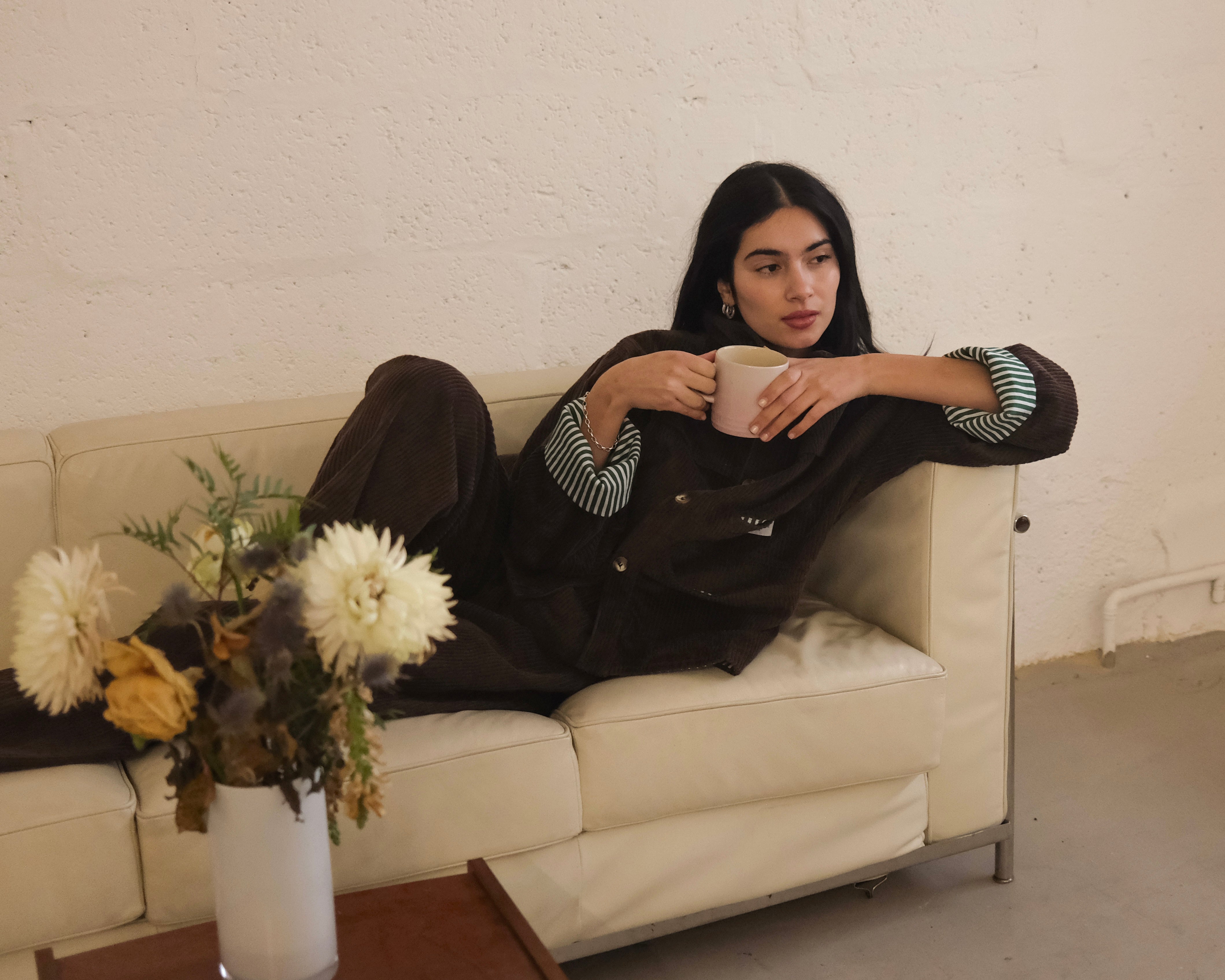 Woman relaxing on a sofa with a mug, wearing the brown corduroy jacket with cuffs rolled to show the Speltham stripe lining