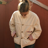 Woman in the cream corduroy jacket against a terracotta wall, hands in the pockets with the striped lining showing at the cuffs