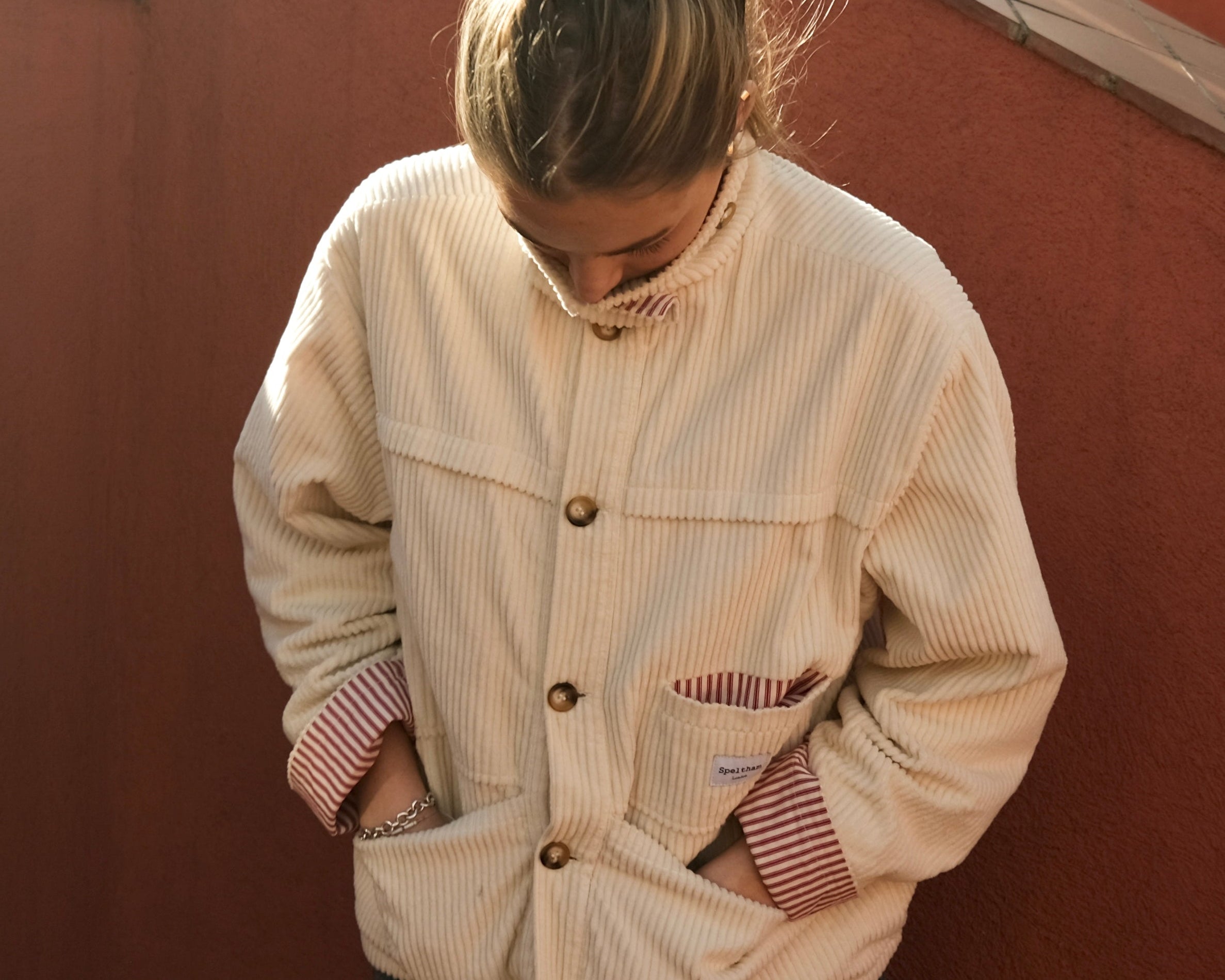 Woman in the cream corduroy jacket against a terracotta wall, hands in the pockets with the striped lining showing at the cuffs